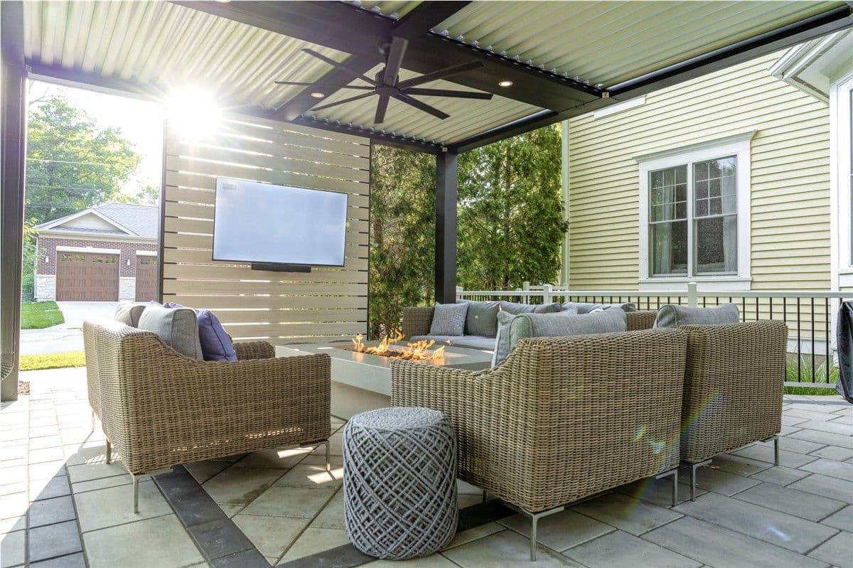 Light wicker furniture sits around a rectangular fire pit under a covered pergola. A ceiling fan hangs above and a TV is mounted on a privacy wall.