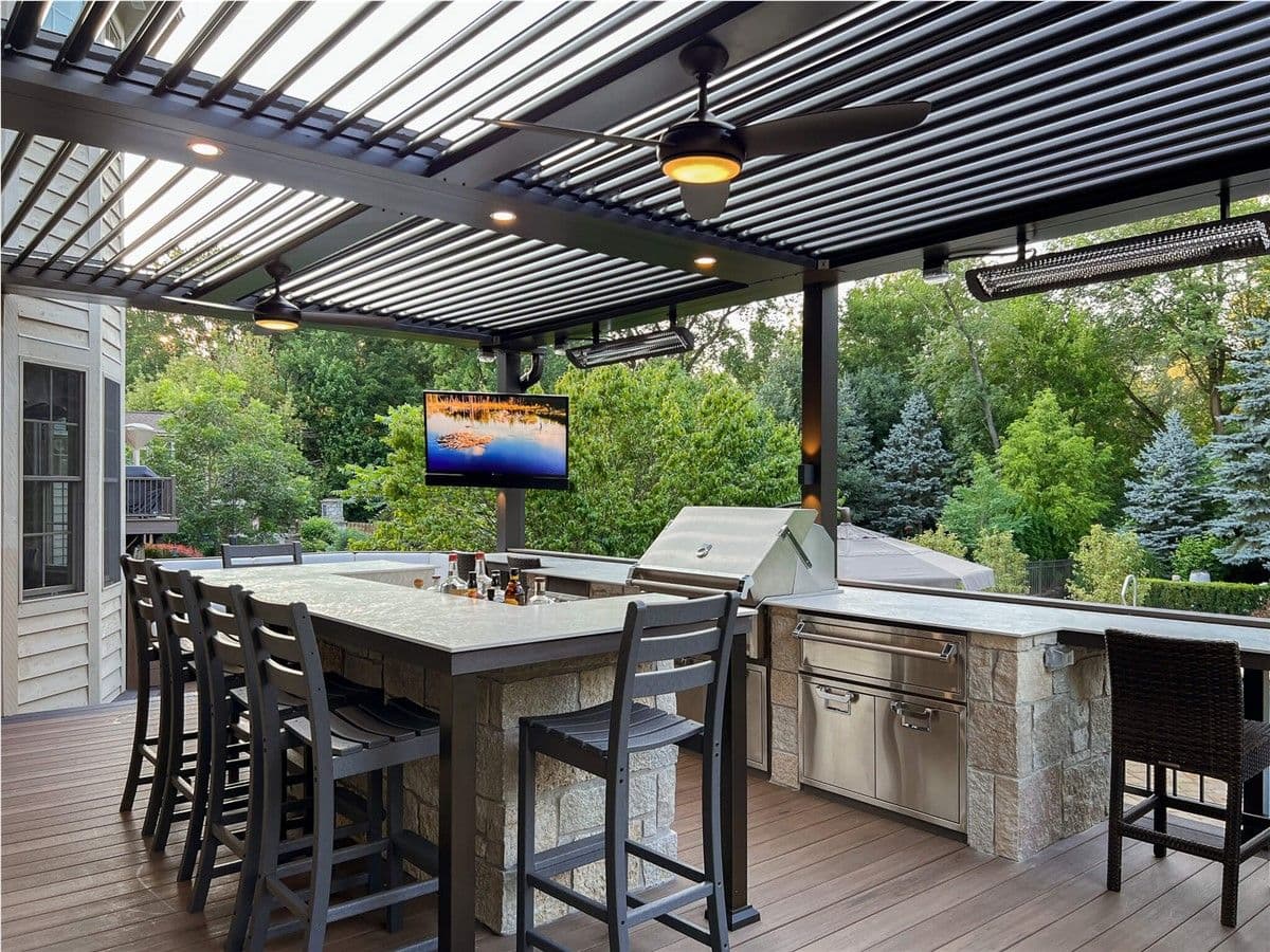 Deck with a dark-colored pergola, built-in stone bar and grill area with stainless steel appliances, bar stools, and a wall-mounted TV, surrounded by trees and landscaping.
