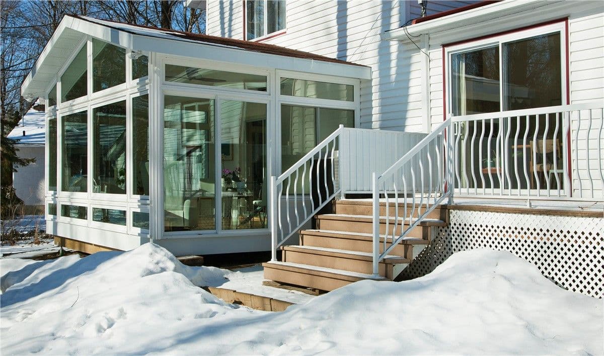 A fully enclosed sunroom with floor to ceiling windows from the outside in winter. Snow is piled on the ground outside.