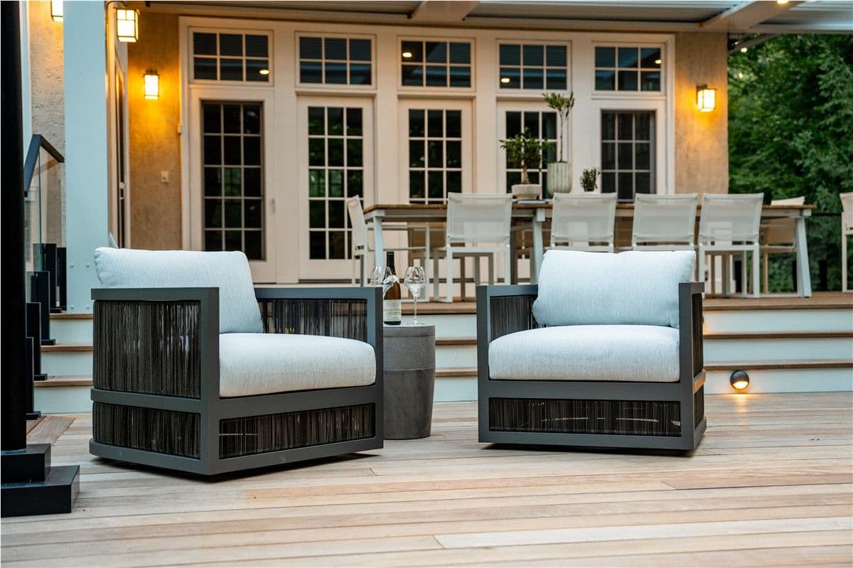 Two dark patio arm chairs with light cushions on a deck. A dining area is visible under a pergola in the background.