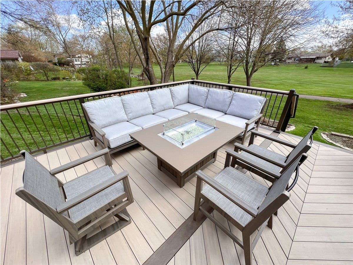 A deck with patio chairs and an l-shaped patio couch around a fire pit overlooks a yard.