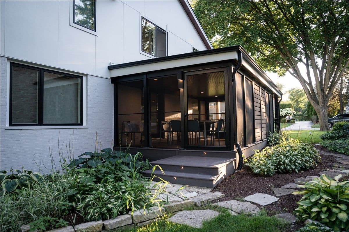 A fully screened-in sunroom off the side of a house. Patio furniture is visible within. A stone path curves around to the right.
