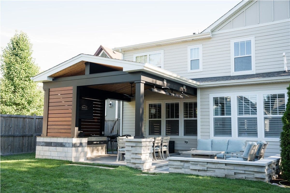 Outdoor living area with a multi-level stone patio, built-in grill, stainless steel appliances, and dark woven furniture, surrounded by green landscaping and trees.