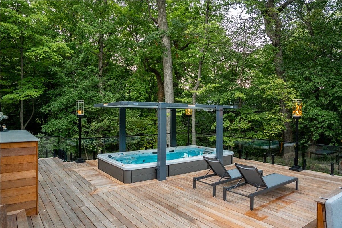 A wood deck with a rectangular, built-in hot tub overlooking a wooded area. Lounge chairs sit to the right.