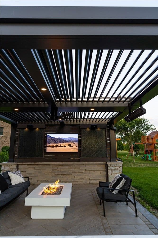 A stone paver patio with a rectangular fire pit beneath a dark-colored Struxure pergola. A TV is mounted on a privacy wall.