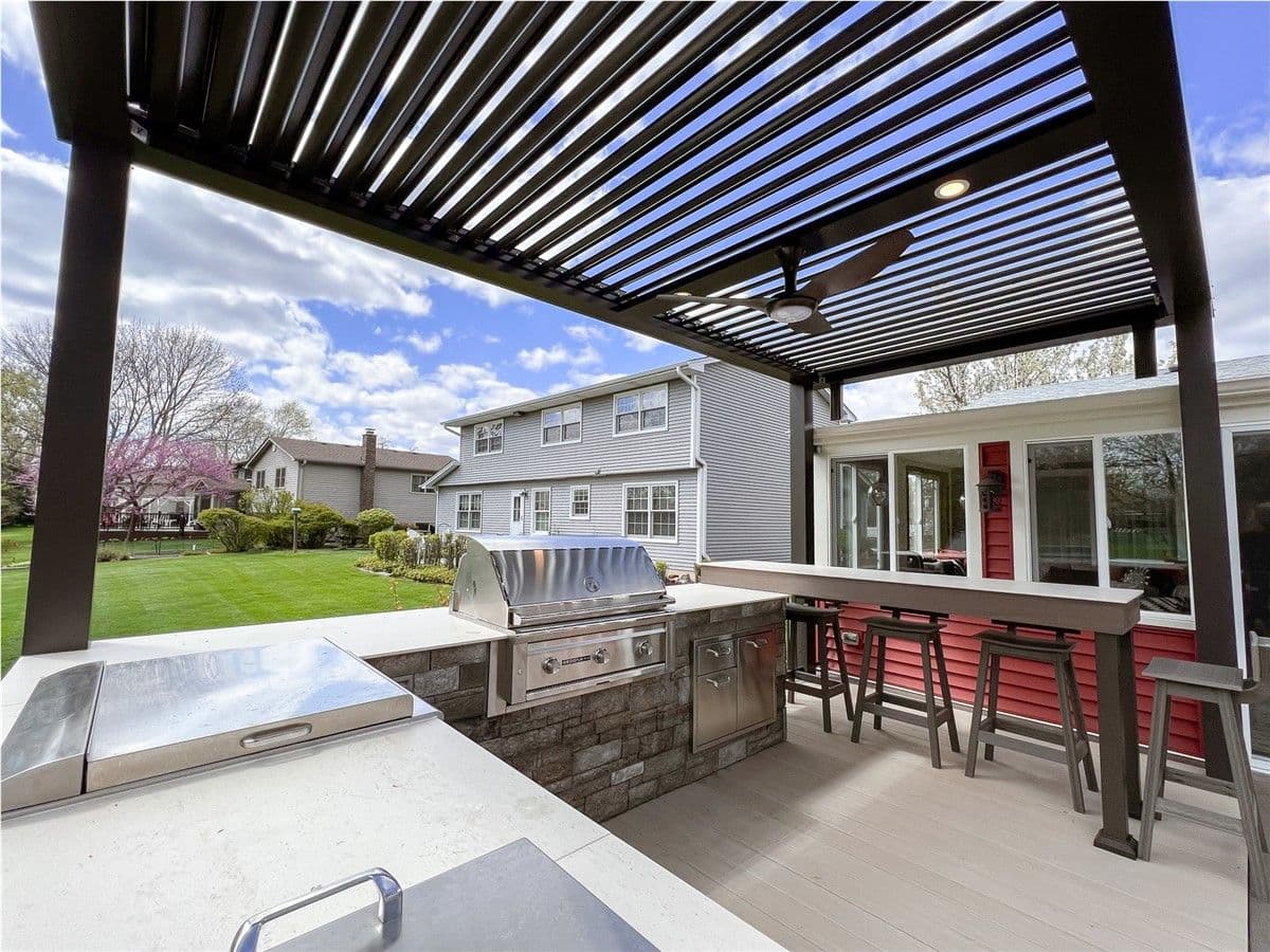 A stone outdoor kitchen and grilling area under a dark-colored pergola. Bar seating is in the background.
