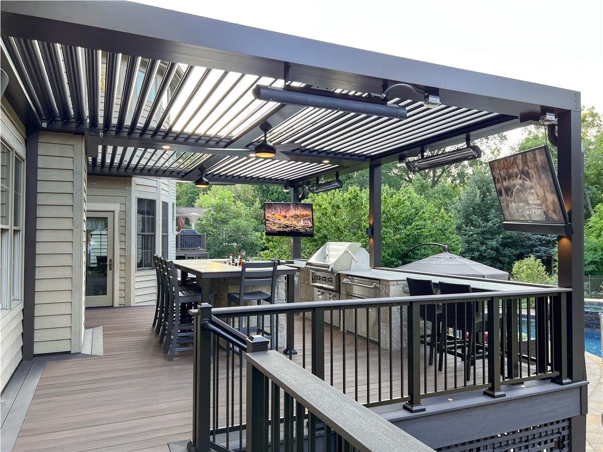 A brown TimberTech deck with a dark-colored pergola, bar and seating, and built-in grilling station. Heating lamps, fans, and mounted TV's hang from the pergola.