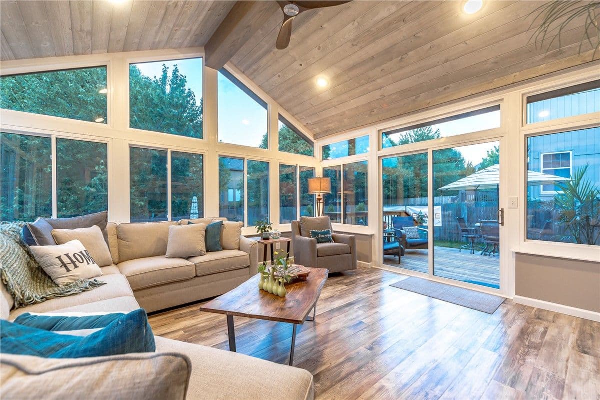 Three-season sunroom with white-framed double-pane windows, a large gray sectional couch, coffee table, and arm chai. A yard with trees and a deck is visible outside.