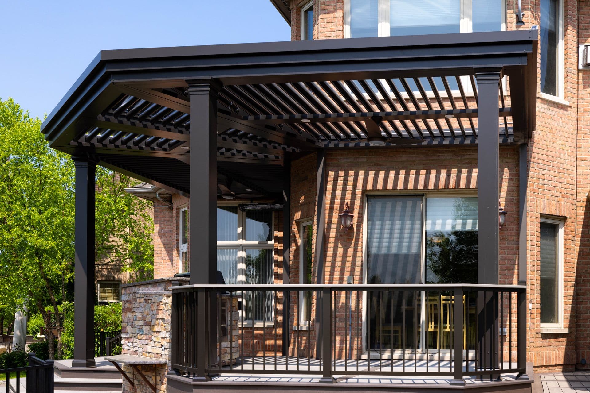 A black custom, wrap-around pergola covers a deck off of a red brick house.